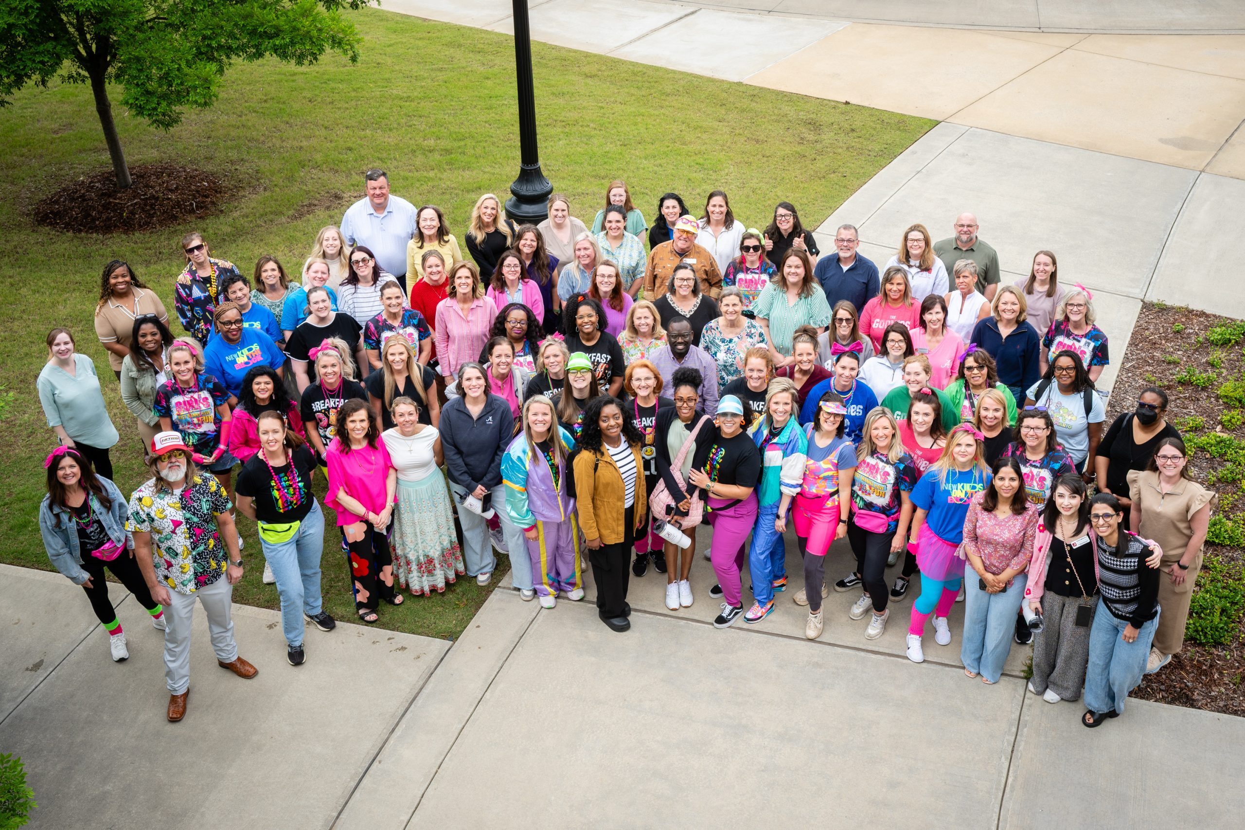 Large group of faculty and staff—approximately 70–80 people—stand closely together on a campus walkway outdoors. The group looks up toward the camera and wears a mix of everyday clothing and bright, themed 1980s‑style outfits with neon colors, graphic T‑shirts, and accessories such as headbands and fanny packs.
